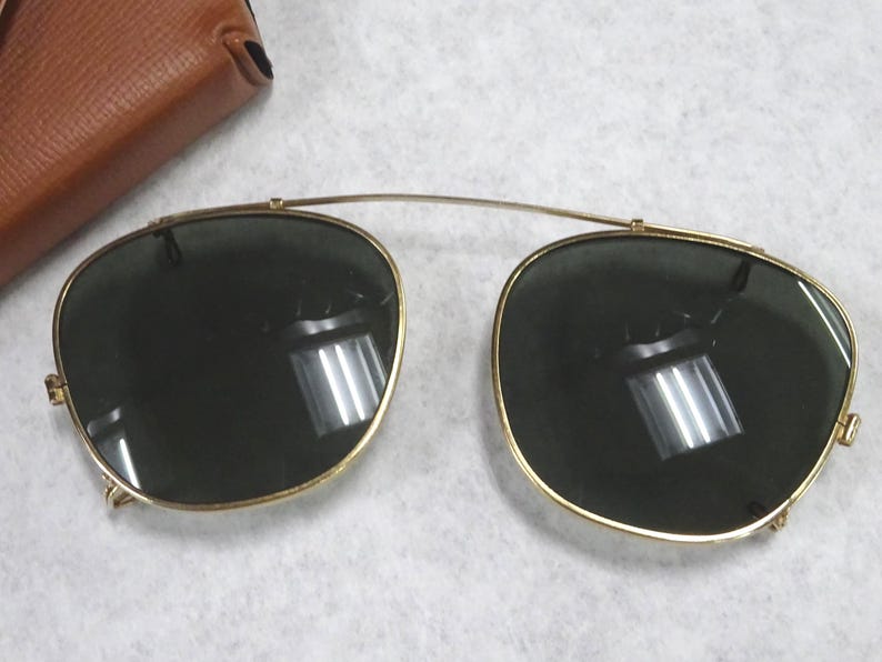 May include: A pair of gold-toned clip-on sunglasses with dark green lenses and a thin metal bridge connecting the two frames. The lenses are rounded and feature a reflective surface. A brown leather-textured case is partially visible in the corner.