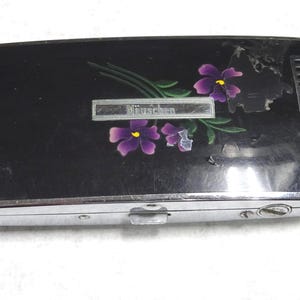 May include: A vintage black cigarette lighter with a silver trim. The lighter features a floral design with purple flowers and green leaves. The word "Müschen" is displayed on a silver rectangular plate.