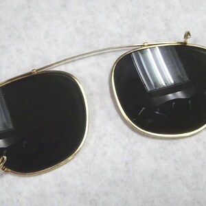 May include: A pair of gold-toned clip-on sunglasses with dark, reflective lenses. The frames feature a thin metal bridge connecting the two rounded rectangular lenses, with small metal clips attached to the edges for securing onto existing eyewear.