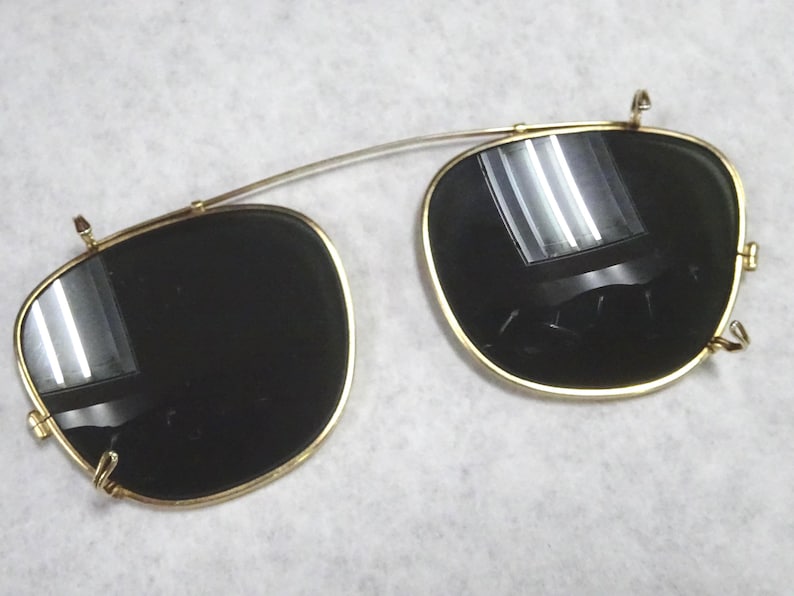 May include: A pair of gold-toned clip-on sunglasses with dark, reflective lenses. The frames feature a thin metal bridge connecting the two rounded rectangular lenses, with small metal clips attached to the edges for securing onto existing eyewear.
