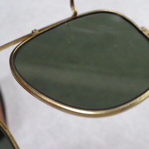 May include: A pair of sunglasses featuring dark green lenses encased in thin, gold-toned metal frames. The design includes a prominent bridge and a structured, rounded lens shape, showcasing a metallic finish on the rims and arms.