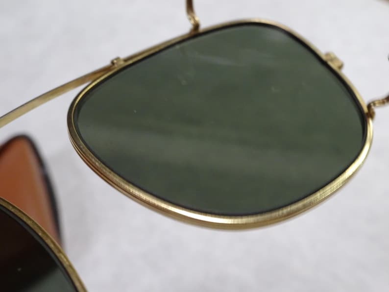 May include: A pair of sunglasses featuring dark green lenses encased in thin, gold-toned metal frames. The design includes a prominent bridge and a structured, rounded lens shape, showcasing a metallic finish on the rims and arms.