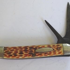 May include: A folding pocket knife with a cream-colored handle featuring a brown spotted pattern. The knife has two dark metal blades extended, silver-toned metal bolsters at each end, and a small silver oval inlay on the handle.