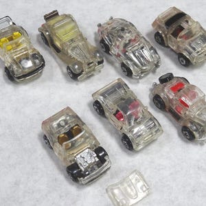 May include: A collection of clear plastic toy cars with black wheels. Some cars have yellow or red interior details. The cars are arranged on a light gray surface. One detached car part is also visible.
