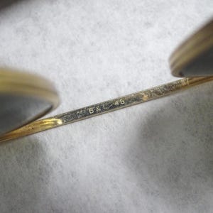 May include: A pair of gold-toned metal eyeglasses featuring a thin bridge engraved with the text B&L 46. The frame includes two circular lens rims, with the metallic structure showing a polished finish and visible markings on the bridge component.
