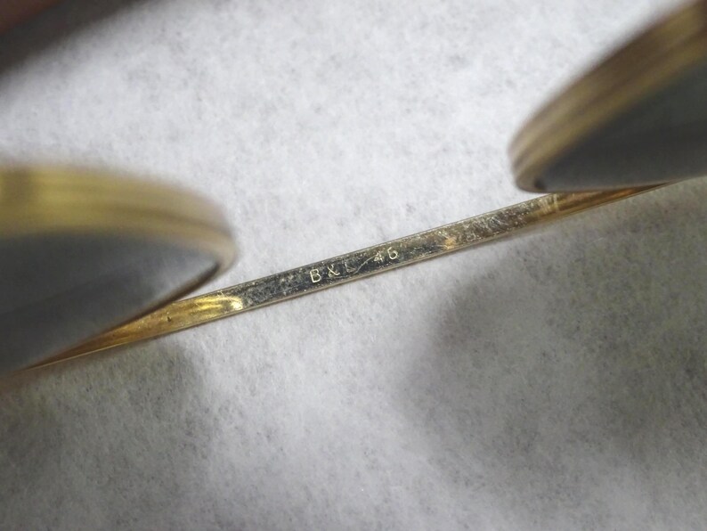 May include: A pair of gold-toned metal eyeglasses featuring a thin bridge engraved with the text B&L 46. The frame includes two circular lens rims, with the metallic structure showing a polished finish and visible markings on the bridge component.