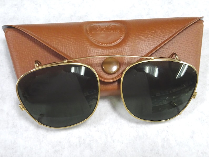 May include: A pair of gold-toned clip-on sunglasses with dark lenses and a thin metal bridge, positioned in front of a tan, textured, triangular-shaped leather carrying case with a snap button closure and a debossed logo on the front flap.