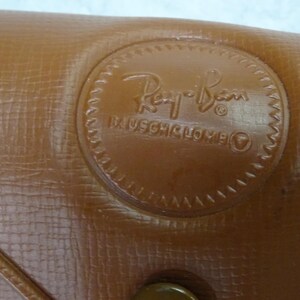 May include: A tan textured case with a circular embossed logo featuring the Ray-Ban script and the text Bausch & Lomb. The surface has a cross-hatched pattern and a visible seam line running diagonally across the lower left corner of the object.