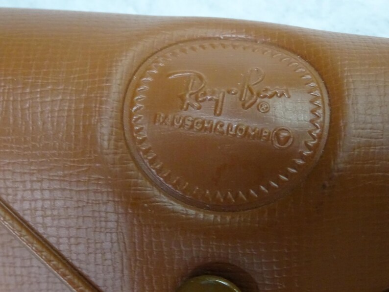 May include: A tan textured case with a circular embossed logo featuring the Ray-Ban script and the text Bausch & Lomb. The surface has a cross-hatched pattern and a visible seam line running diagonally across the lower left corner of the object.