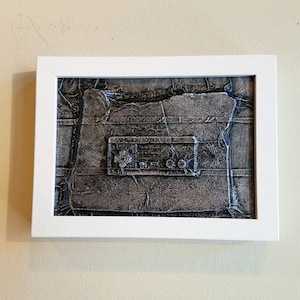May include: A white framed picture of a textured grey and black image of a vintage radio. The radio has a rectangular shape with a textured surface and a small rectangular window in the centre.