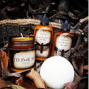 May include: A set of three Halloween-themed bath and body products. A brown glass candle with the text "I'D F**K ME" and a black moth graphic. A brown glass bottle of lotion with the text "It rubs the lotion on the skin" and a black moth graphic. A brown glass bottle of lotion with the text "...Or else it gets the hose again" and a black moth graphic. A white bath bomb.