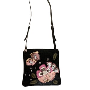 以下が含まれることがあります： 長い調節可能なストラップが付いた黒いスエードのクロスボディバッグ。バッグには、ラインストーンと蝶のデザインが施されたピンクとグリーンのフラワーアップリケが施されています。