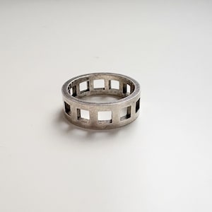 May include: A silver ring with a geometric design. The ring features a series of square cutouts around the band. The ring is made of a shiny, metallic material and is set against a white background. The ring is a piece of jewelry.