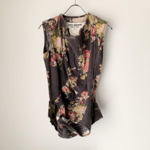 Puede incluir: Un top sin mangas con estampado floral con una etiqueta de Junya Watanabe. El top es de color gris oscuro con flores rosas, blancas y amarillas. El top tiene un diseño único con una cintura ajustada y un escote drapeado.