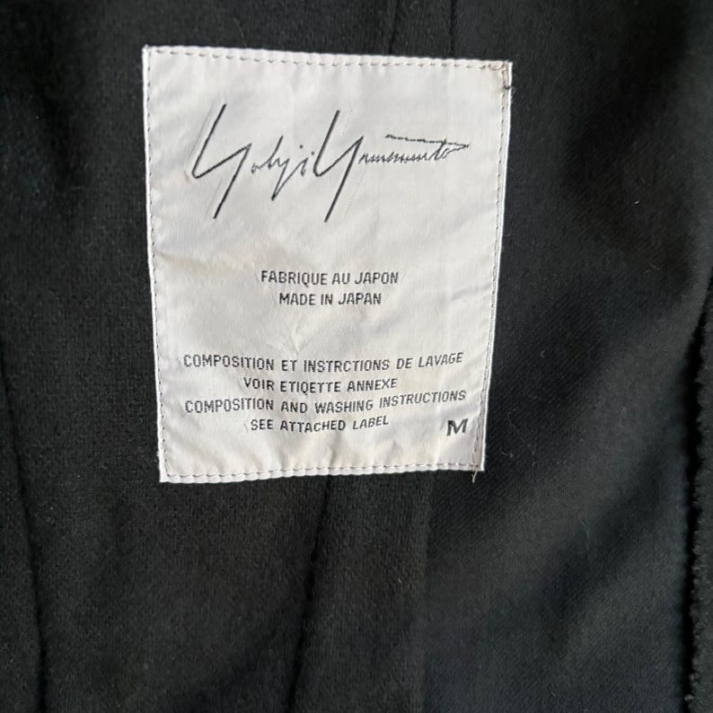 Puede incluir: Primer plano de una prenda negra con una etiqueta blanca. La etiqueta incluye el texto "FABRIQUE AU JAPON MADE IN JAPAN" e instrucciones de lavado en franc&eacute;s e ingl&eacute;s. La talla est&aacute; marcada como "M".