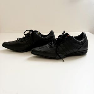 May include: A pair of black leather sneakers featuring a low-top silhouette, rounded toes, and black laces. The shoes have a smooth finish with tonal stitching details, a padded collar, and a flat black rubber sole with a textured grip pattern.