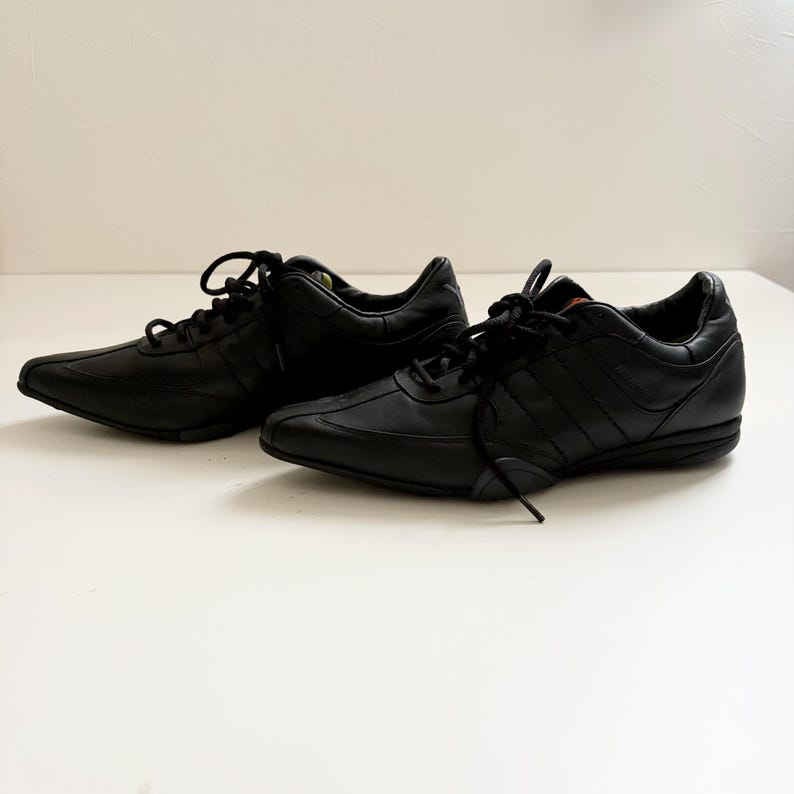 May include: A pair of black leather sneakers featuring a low-top silhouette, rounded toes, and black laces. The shoes have a smooth finish with tonal stitching details, a padded collar, and a flat black rubber sole with a textured grip pattern.