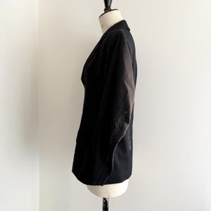 Puede incluir: Un blazer negro exhibido en un maniqu&iacute;. El blazer tiene un dise&ntilde;o cl&aacute;sico con solapa de muesca y cierre de dos botones. Las mangas presentan costuras blancas decorativas. La prenda est&aacute; hecha de una tela oscura.
