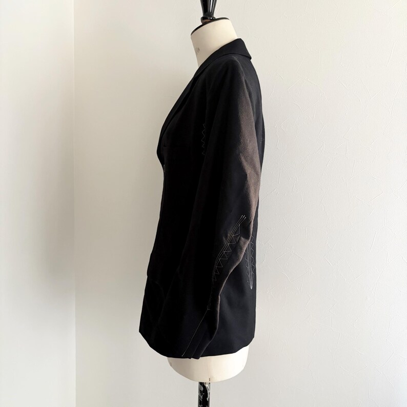 Puede incluir: Un blazer negro exhibido en un maniqu&iacute;. El blazer tiene un dise&ntilde;o cl&aacute;sico con solapa de muesca y cierre de dos botones. Las mangas presentan costuras blancas decorativas. La prenda est&aacute; hecha de una tela oscura.