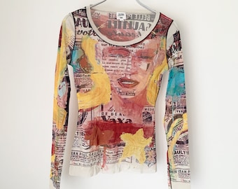 May include: A long-sleeved, sheer, white mesh top with a colorful print of a woman's face, text, and abstract shapes. The print includes the text "AUAJIHUA", "vota tu", "sur To", "paraitrez", "MEDE", "DE BEA", "Vaya", "JEAN PAUL GAULIER", "AULTI", and "SETS JEAN P".
