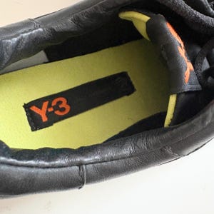 May include: A black sneaker featuring a lime green insole with a black rectangular patch displaying the orange Y-3 logo. The interior collar is lined with lime green fabric, and the tongue features an orange Y-3 logo on a black background.