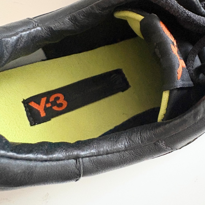 May include: A black sneaker featuring a lime green insole with a black rectangular patch displaying the orange Y-3 logo. The interior collar is lined with lime green fabric, and the tongue features an orange Y-3 logo on a black background.