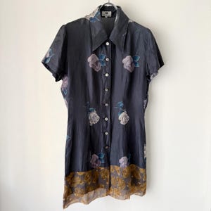 May include: A dark grey button-down shirt dress with short sleeves and a floral pattern. The dress features white buttons, a collar, and a decorative floral border at the hem. The floral pattern includes roses in shades of purple, blue, and white.