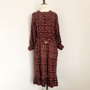 Puede incluir: Un vestido midi marrón con un patrón geométrico de cuadrados rosas, rojos y beige. El vestido tiene manga larga, un frente abotonado y un cinturón con una hebilla dorada. El vestido tiene un dobladillo plisado.