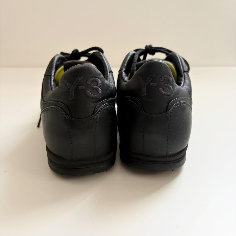 May include: A pair of black leather sneakers featuring a low-top silhouette with black laces and a lime green interior lining. The heel counter of each shoe is embossed with the Y-3 logo in a matching black tone, showcasing a smooth finish.