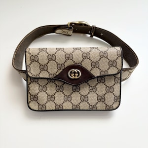 May include: A brown leather belt with a gold buckle attached to a beige and brown canvas fanny pack with a brown leather flap and a gold GG logo.