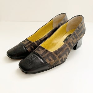 May include: A pair of vintage brown and black square-toe pumps. The shoes feature a repeating brown pattern on a tan background, with black leather toe caps. The heels are a dark brown color, and the insoles are yellow.