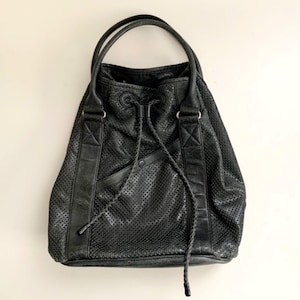 May include: Black leather tote bag with a perforated design. The bag has two handles and a drawstring closure. The bag has a small pocket on the front and is made of a soft, supple leather.