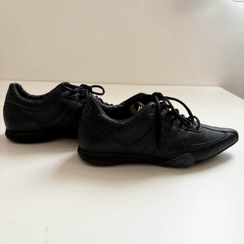 May include: A pair of black low-top sneakers featuring a smooth leather upper, tonal stitching, and black laces. The shoes have a rounded toe, a padded collar, and a low-profile black rubber sole. The design includes subtle paneling details.