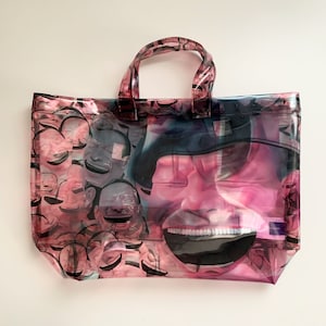 May include: A clear plastic tote bag with a pink and black print of a smiling face. The face is surrounded by smaller smiling faces.