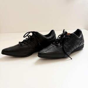 May include: A pair of black low-top sneakers featuring a smooth leather upper, tonal stitching, and black laces. The shoes have a pointed toe design, a low-profile rubber sole, and a small orange logo detail visible on the tongue of the right shoe.