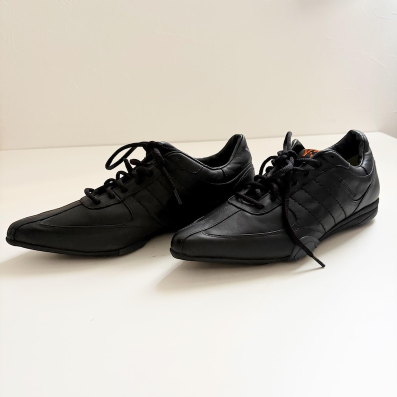 May include: A pair of black low-top sneakers featuring a smooth leather upper, tonal stitching, and black laces. The shoes have a pointed toe design, a low-profile rubber sole, and a small orange logo detail visible on the tongue of the right shoe.