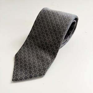 May include: A dark gray tie with a repeating pattern of the letter "G" in a lighter gray. The tie is made of a smooth fabric and is neatly folded, showcasing its design and texture.