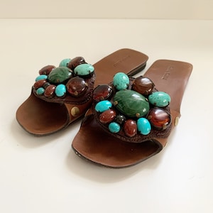 May include: Brown leather sandals with a large cluster of turquoise, green, and brown stones. The sandals have a simple design and are perfect for summer.