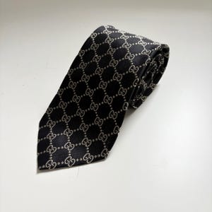 May include: Black necktie with a repeating pattern of interlocking G logos in a lighter color. The tie is made of a smooth fabric and is displayed on a white surface.