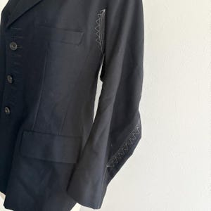 Puede incluir: Un blazer negro con un corte entallado. El blazer presenta una solapa con muescas, un cierre de bot&oacute;n sencillo y un peque&ntilde;o bolsillo en el pecho. Las costuras blancas acent&uacute;an las costuras de las mangas. La chaqueta est&aacute; hecha de una tela oscura y lisa.
