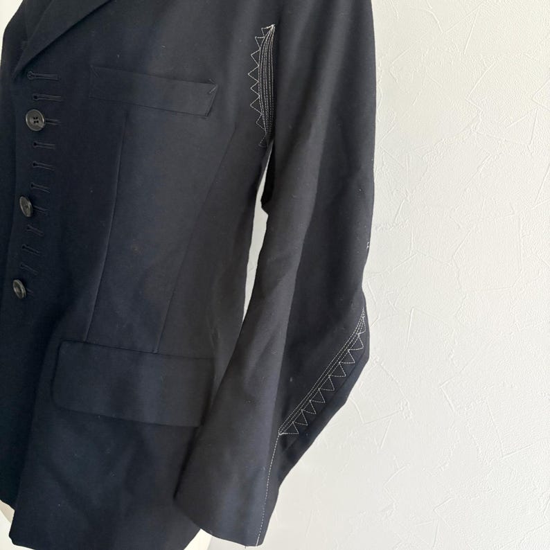 Puede incluir: Un blazer negro con un corte entallado. El blazer presenta una solapa con muescas, un cierre de bot&oacute;n sencillo y un peque&ntilde;o bolsillo en el pecho. Las costuras blancas acent&uacute;an las costuras de las mangas. La chaqueta est&aacute; hecha de una tela oscura y lisa.