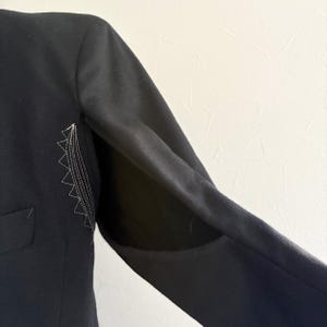 Puede incluir: Manga de blazer negra con un dise&ntilde;o cosido blanco en la parte superior del brazo. La manga est&aacute; en &aacute;ngulo, mostrando el forro interior. El blazer tiene un bolsillo en la parte delantera.