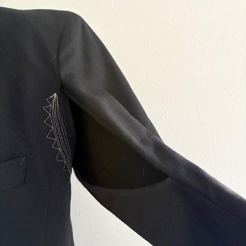 Puede incluir: Manga de blazer negra con un dise&ntilde;o cosido blanco en la parte superior del brazo. La manga est&aacute; en &aacute;ngulo, mostrando el forro interior. El blazer tiene un bolsillo en la parte delantera.