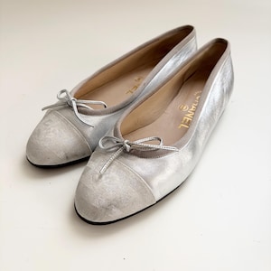 May include: A pair of silver ballet flats featuring a metallic finish, a rounded toe with a textured fabric cap, and a small decorative bow on the front. The interior lining is beige with the gold Chanel logo printed on the heel of the insole.
