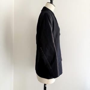 Puede incluir: Blazer negro con solapa de muesca y corte entallado. La chaqueta presenta costuras decorativas a lo largo de la costura lateral. El blazer se exhibe en un maniqu&iacute; sobre un fondo blanco.