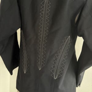 Puede incluir: Chaqueta negra con detalles de costura en zigzag blancos en la espalda. La chaqueta tiene mangas largas y un corte entallado. La costura crea un patr&oacute;n decorativo, agregando un elemento de dise&ntilde;o &uacute;nico a la prenda.
