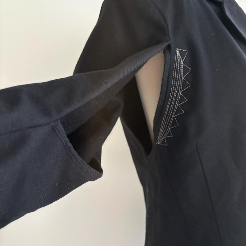 Puede incluir: Primer plano de una blazer negra con un corte entallado. La manga presenta un dise&ntilde;o &uacute;nico con una abertura abierta y costuras decorativas en un color claro contrastante. La tela de la blazer parece ser un material oscuro y resistente.
