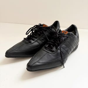 May include: A pair of black leather sneakers featuring a pointed toe design, black laces, and the orange Y-3 logo printed on the tongue. The shoes have a low-top silhouette with stitched paneling details along the sides and a black rubber sole.