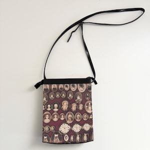 May include: A rectangular crossbody bag with a black strap and top. The bag's main body features a collage of vintage portraits and clocks on a burgundy background. The strap is thin and black, and the bag is hanging against a white background.