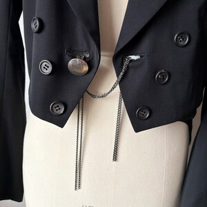 May include: A black cropped blazer with a silver chain and a circular pendant. The blazer has multiple buttons and a pointed collar.